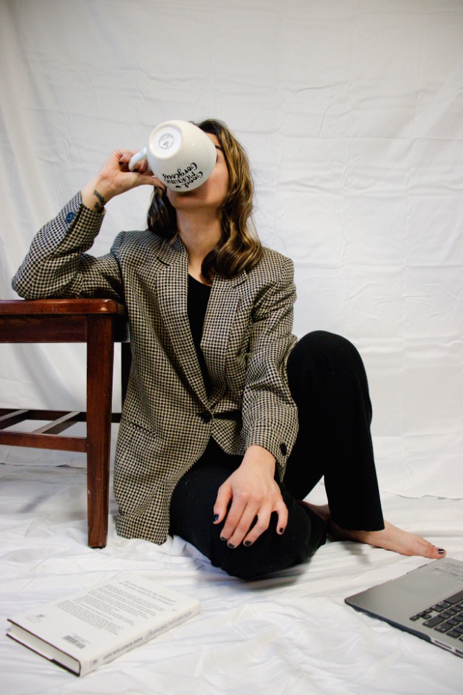 Sylvana chugs her coffee with a joyful grin, sitting casually with one arm on the chair, ready to connect with others on her computer.